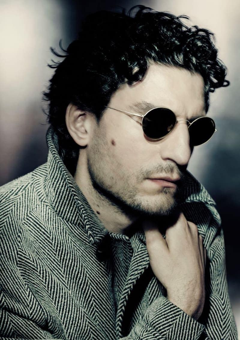 Portrait of a man with dark curly hair wearing sunglasses and a grey herringbone coat, posing in front of a blurred backdrop.