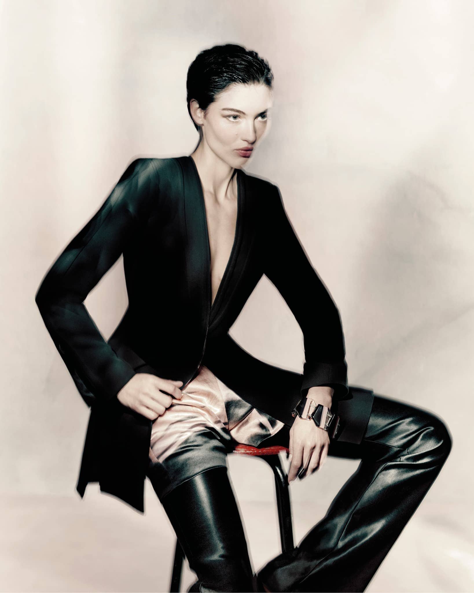 A fashion shot of a woman sitting on a stool wearing a black jacket, metallic shorts, and shiny black thigh-high boots in a studio.