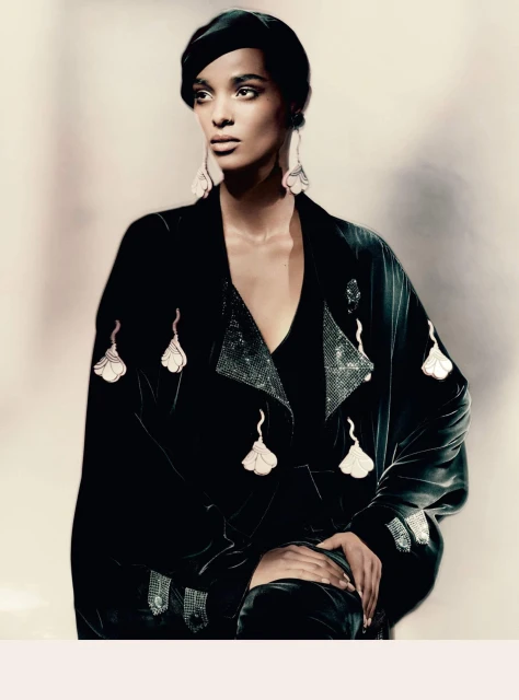A black woman with dark hair wearing a black velvet coat and black hat with hanging floral ornaments and drop earrings in a studio.