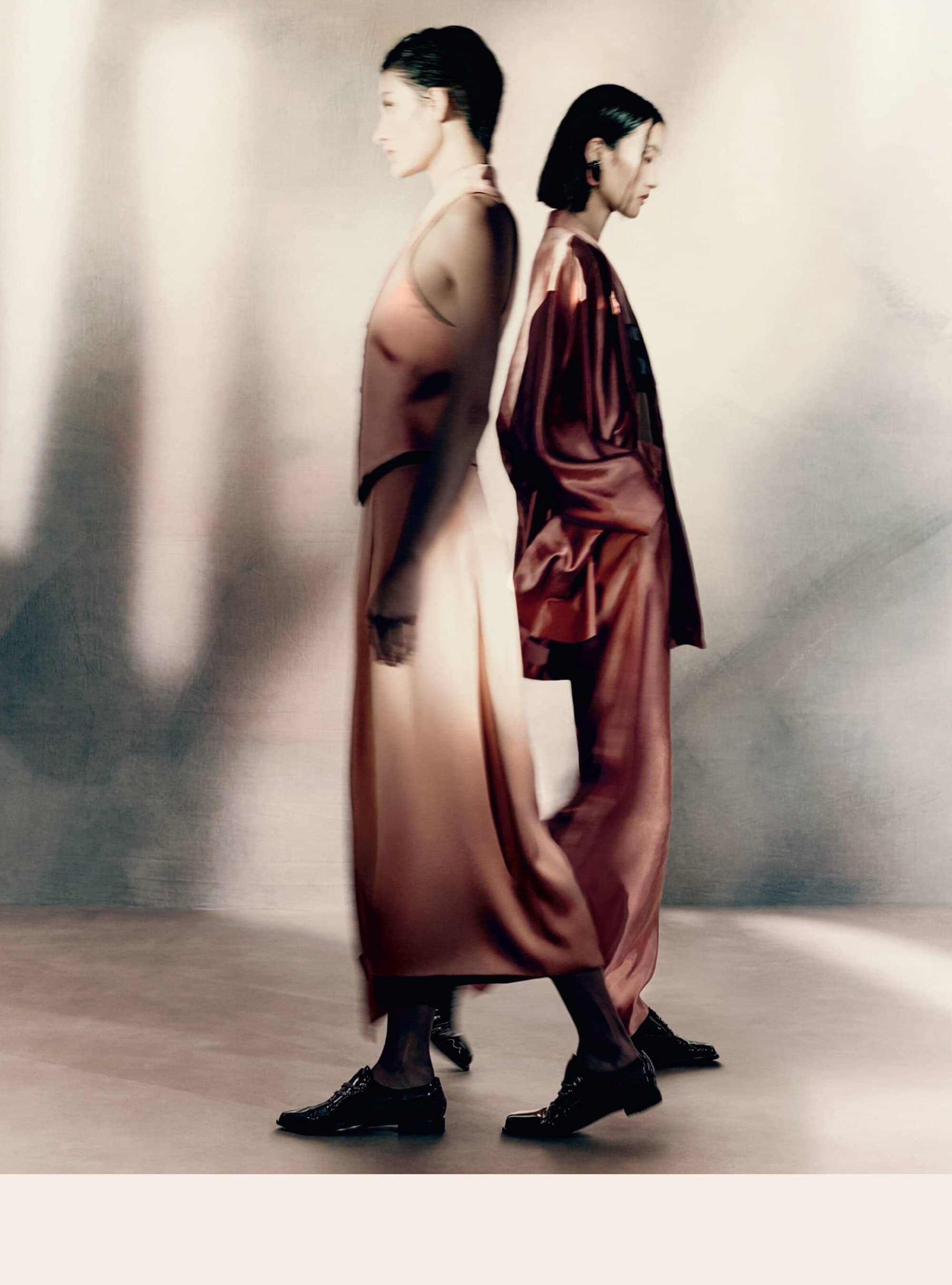Two fashion models in satin outfits stand back-to-back, one in peach and the other in burgundy, with blurred motion effect on a neutral background.