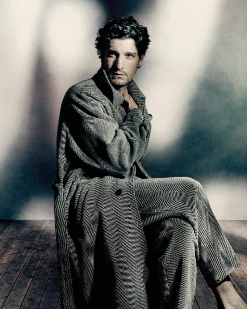 Adult male poses in a herringbone pattern coat and pants, seated on a wooden floor, against a gradient gray background with soft lighting.