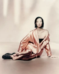 Model Lina Zhang seated on a studio floor wearing a glossy rose-gold satin suit and black shoes for Giorgio Armani.