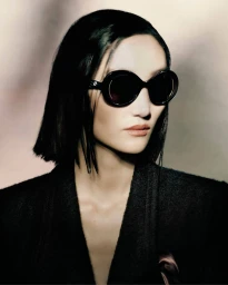 Portrait of a woman wearing dark round sunglasses and a dark blazer against a soft gradient background.