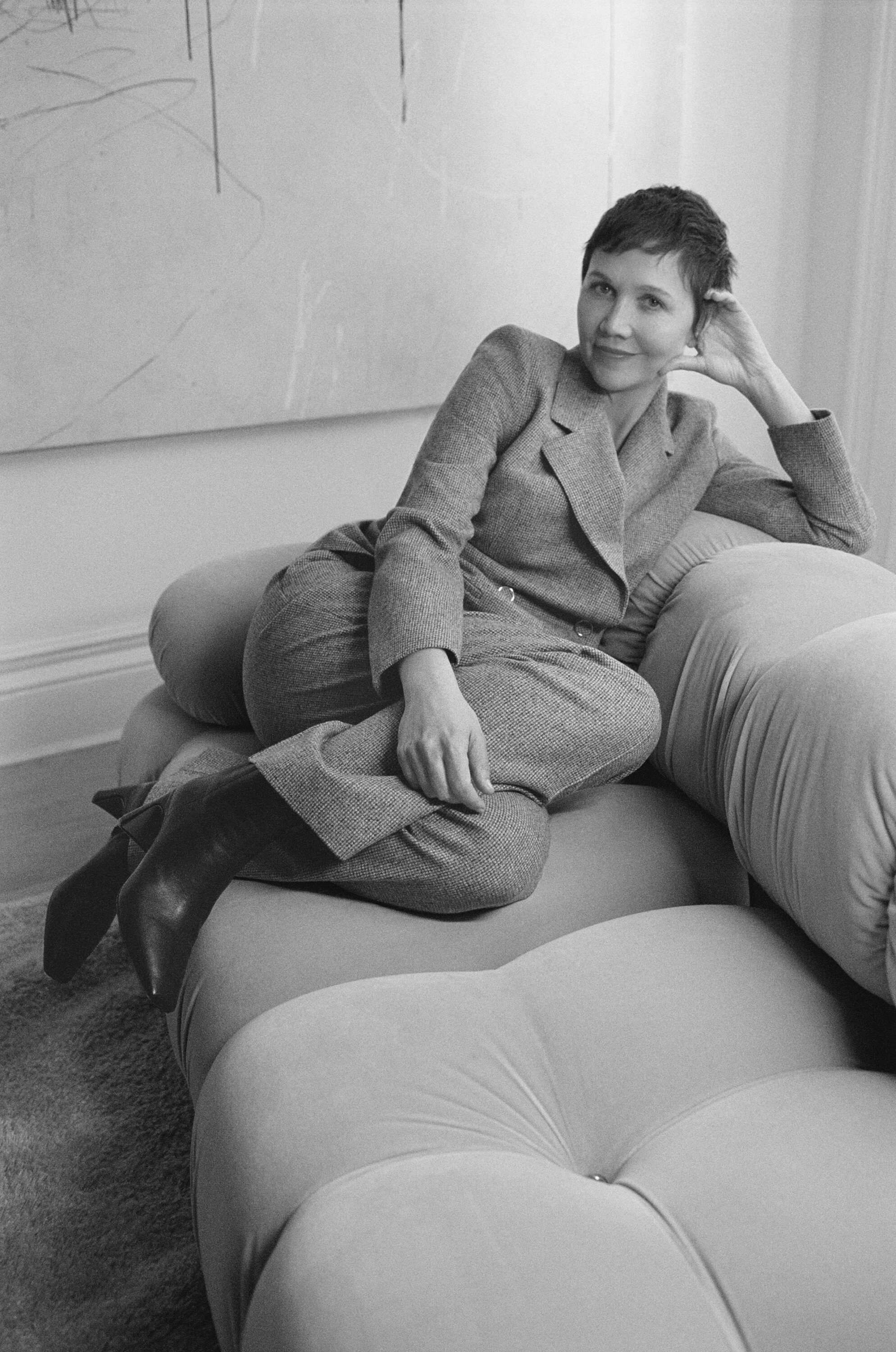 Maggie Gyllenhaal reclining on a modular sofa wearing a tailored grey suit and black boots in black and white.