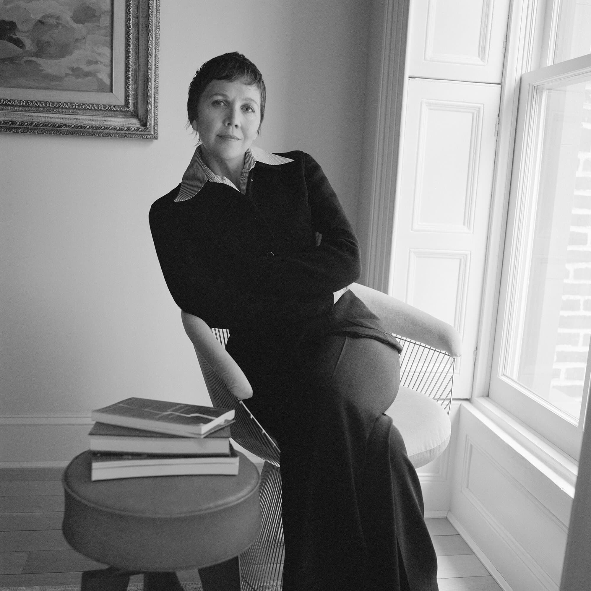 Monochrome portrait of a stylish woman with short hair sitting in a chair near a window, next to a small table with books.
