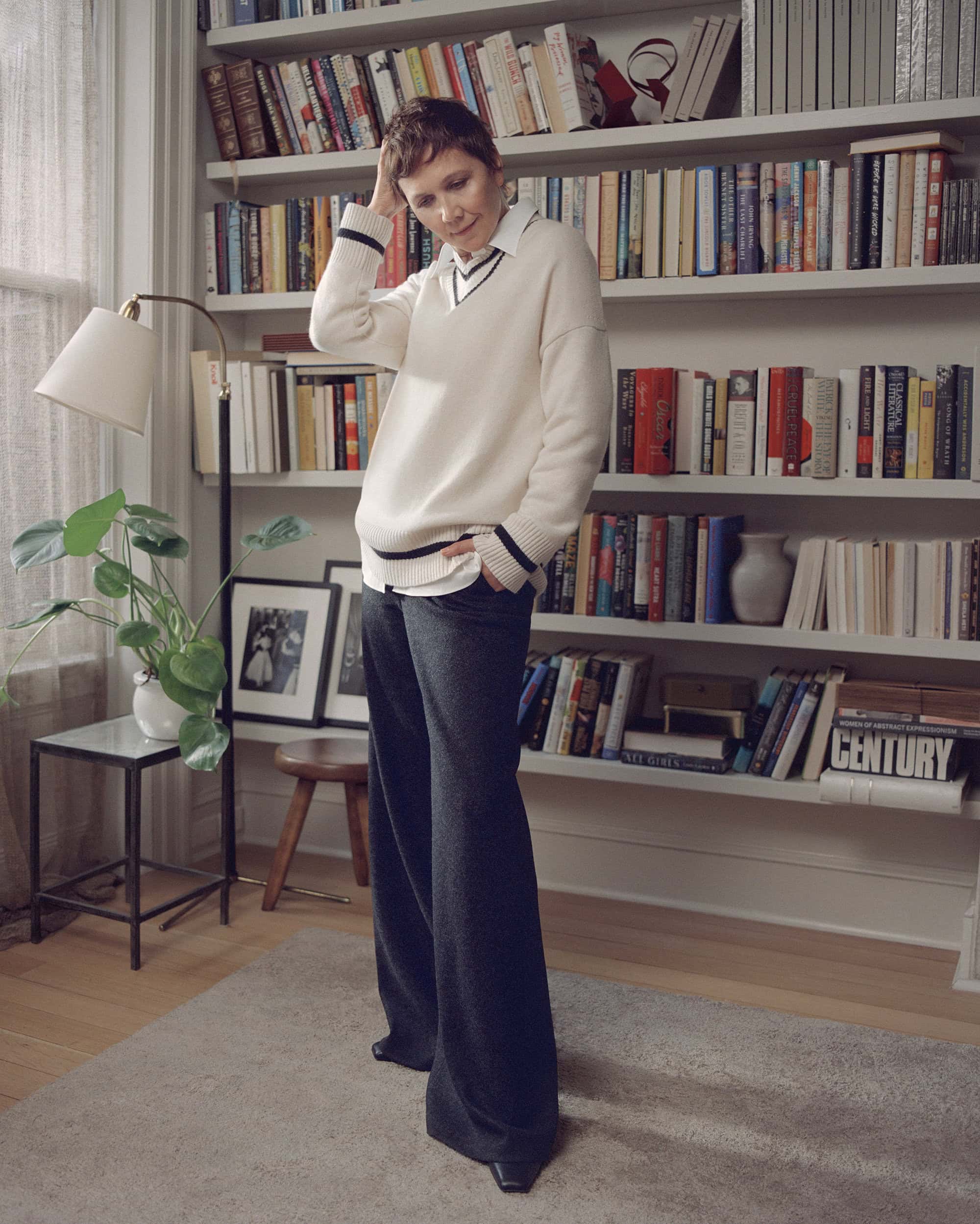 A mature woman with short hair stands indoors in a relaxed pose, wearing a cream sweater, wide-legged pants, and black shoes, next to a bookshelf.