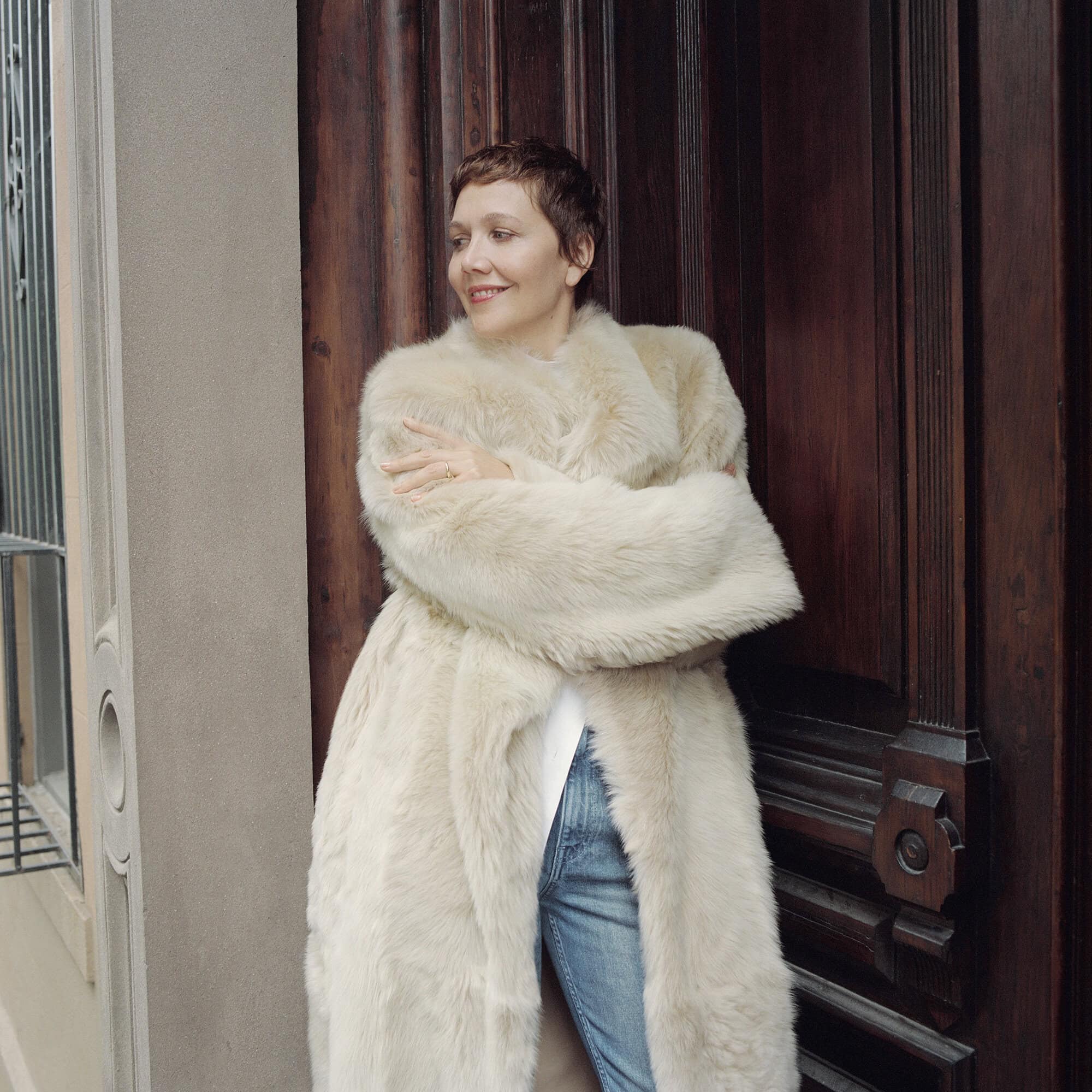 Adult female with short red hair in a full-length beige fur coat posing outdoors in front of a dark brown carved wooden door.