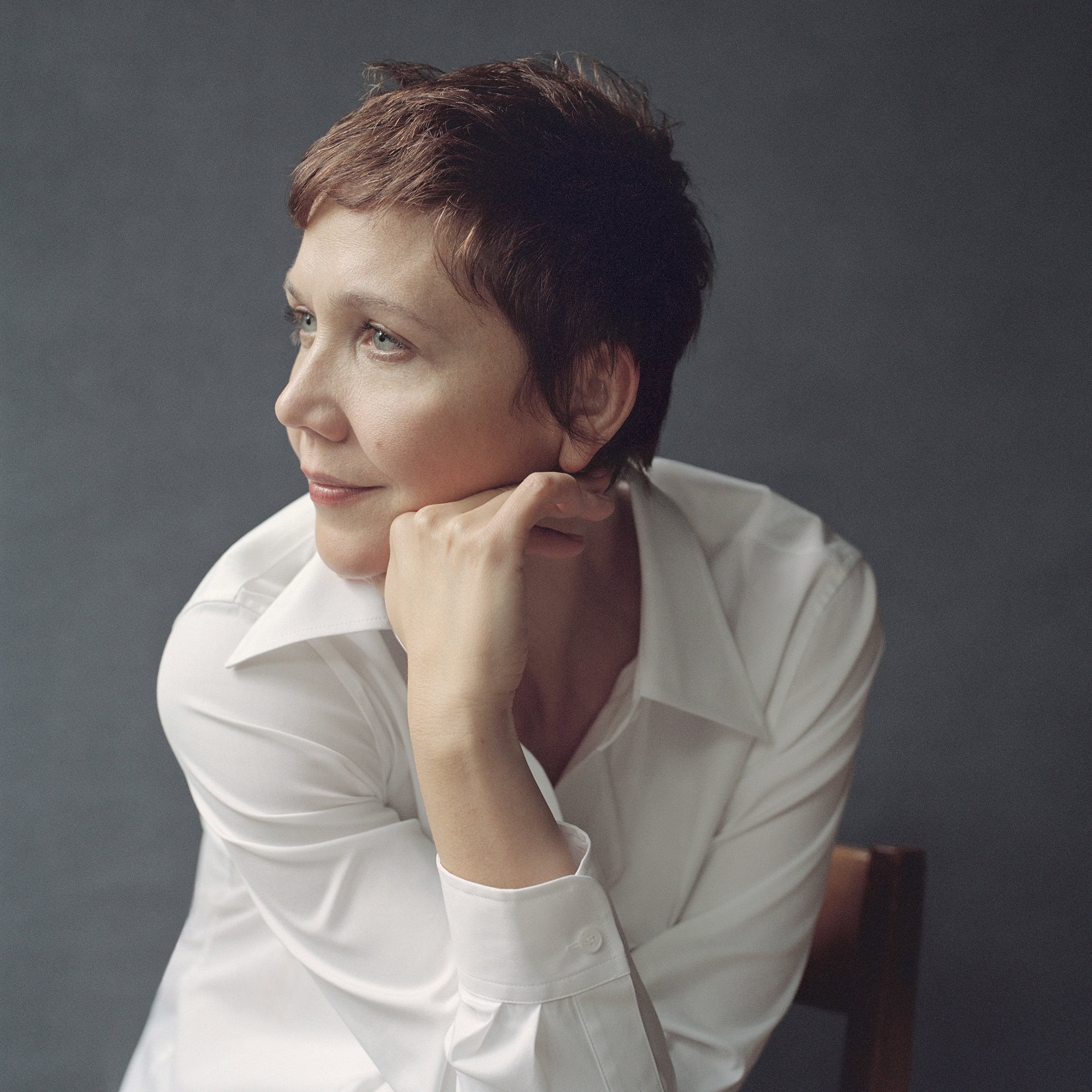 Maggie Gyllenhaal resting her chin on her hand wearing a crisp white button-down shirt against a dark grey studio backdrop.