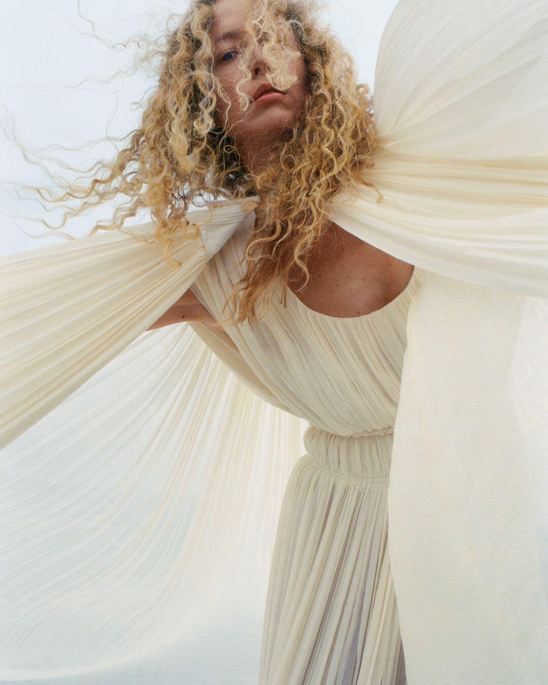 A woman with blonde curly hair is wearing an off-white pleated dress and shawl against a bright, washed-out sky background in a fashion editorial.