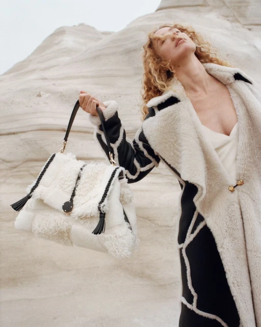Fashion shot of a woman in a shearling coat holding a shearling purse, posing in front of a light-colored rock formation.