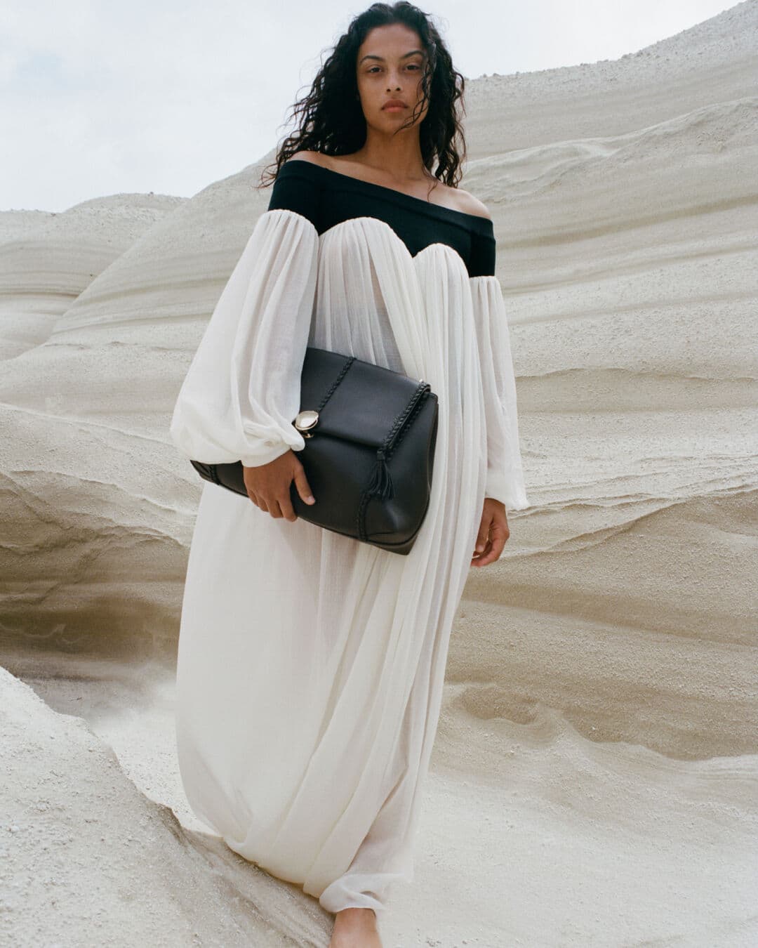 Adult female in long white off-shoulder dress and black handbag poses against a stark, bright, desert-like background with unique rock formations.