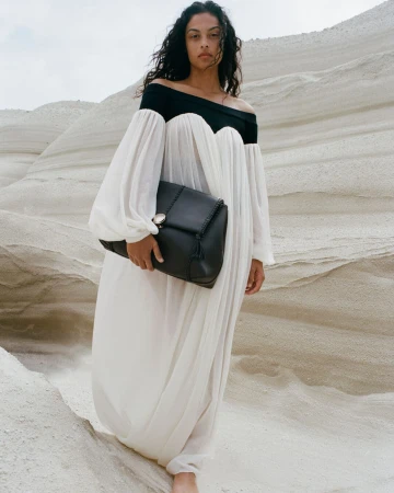 Adult female in long white off-shoulder dress and black handbag poses against a stark, bright, desert-like background with unique rock formations.