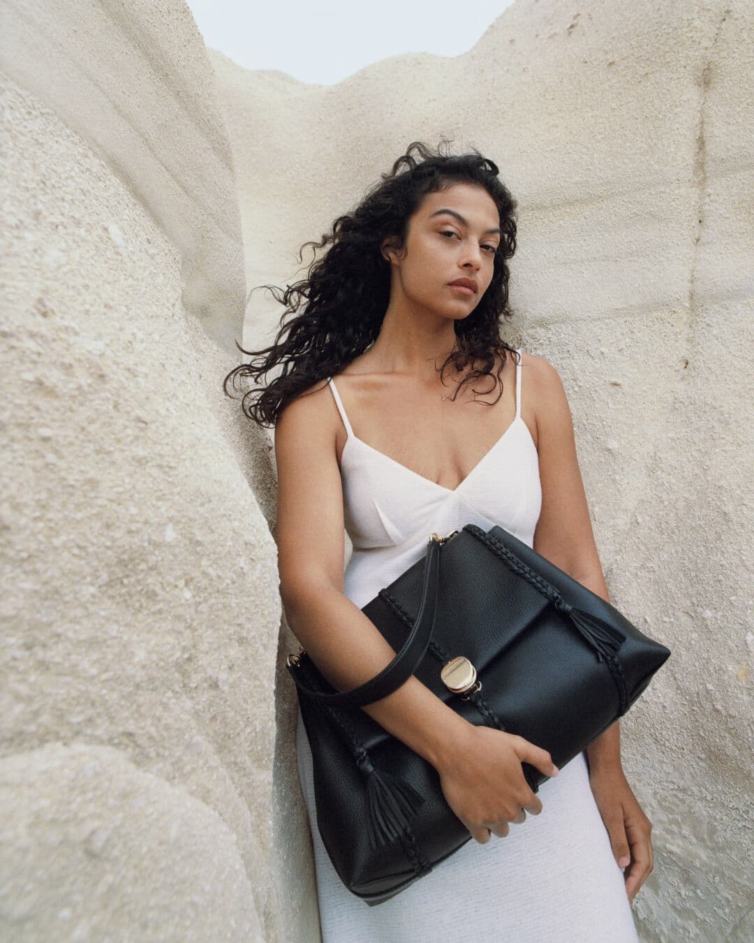 Brunette woman in white dress poses with large black leather satchel handbag against neutral geological background, fashion-forward product shot.