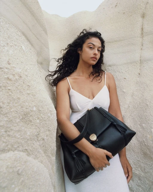 Brunette woman in white dress poses with large black leather satchel handbag against neutral geological background, fashion-forward product shot.