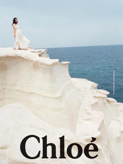 A woman in a white dress stands on a white cliff overlooking the ocean under a pale sky, promoting Chloé fashion in an outdoor setting.