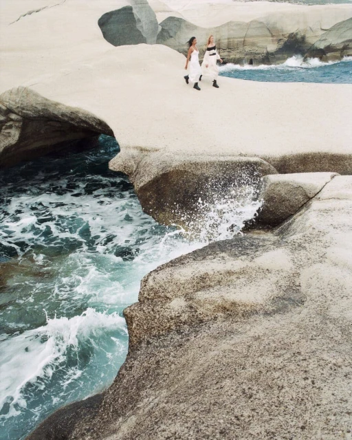 Two adult females walk along a white rock formation near ocean waves with a blue-sky background; sunlit, outdoor fashion photography.