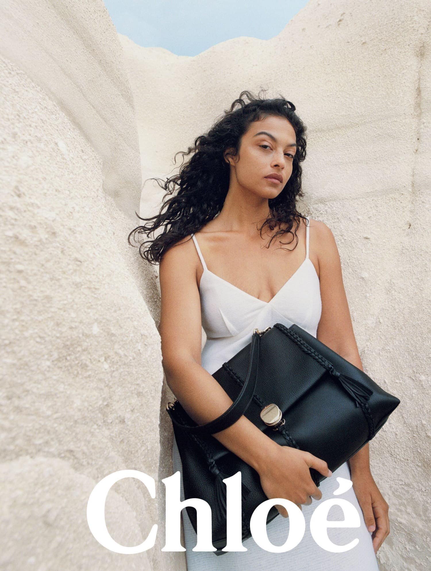 A woman poses with a black Chloe purse in a bright outdoor setting with white rock formations, showcasing fashion and style.
