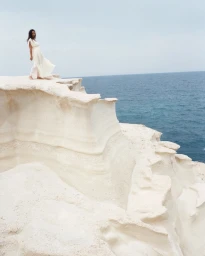 Model Devyn Garcia wearing a flowing white maxi dress atop sculptural white cliffs overlooking the sea in Milos, Greece.