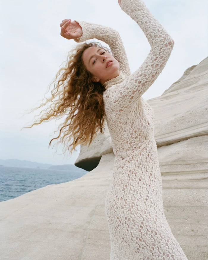 Model with long curly hair wearing a cream lace dress posing against white rocky cliffs near the ocean.