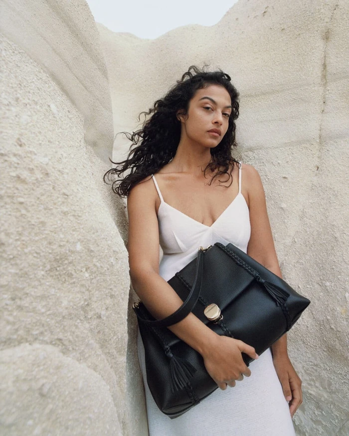 Devyn Garcia in a white slip dress holding a black leather handbag against pale rock formations.