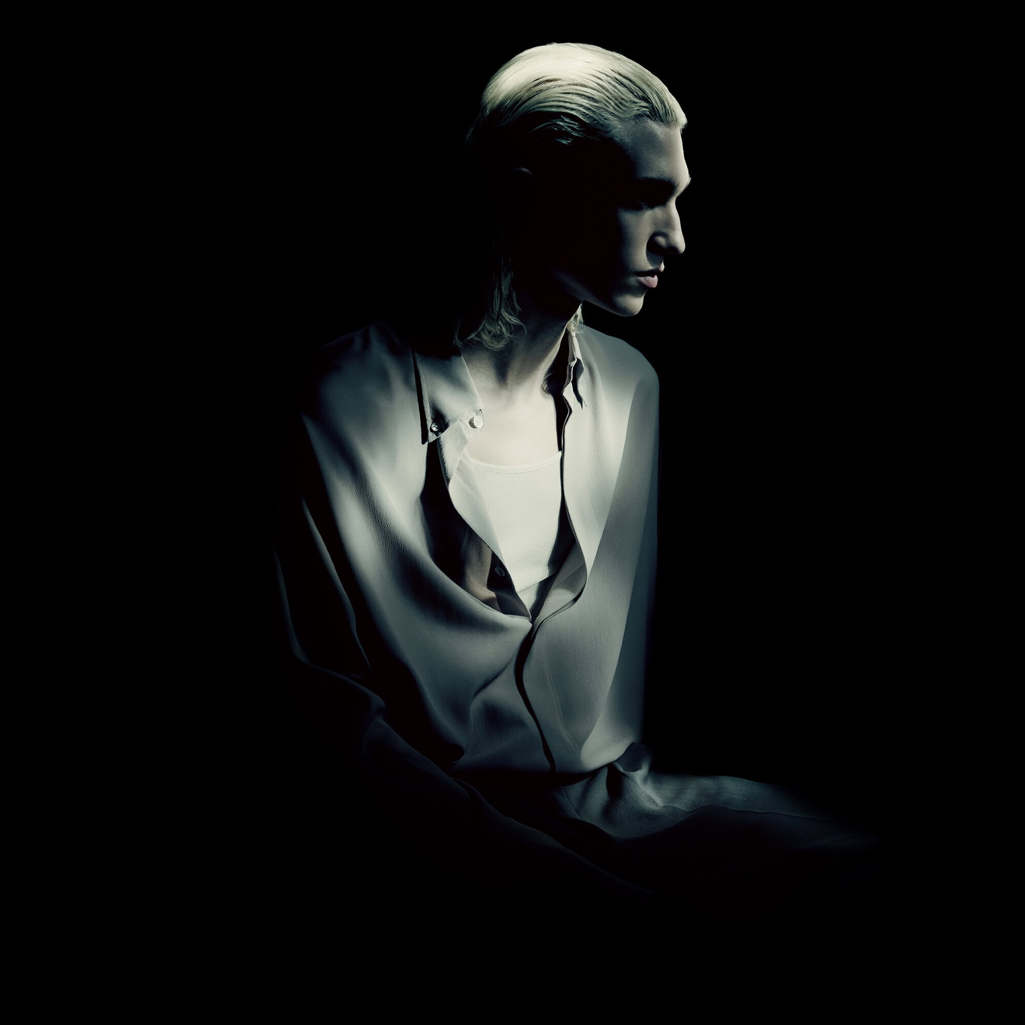 Portrait of a young adult with fair skin and blonde hair, wearing a loosely fitted shirt against a dark, shadowed background.