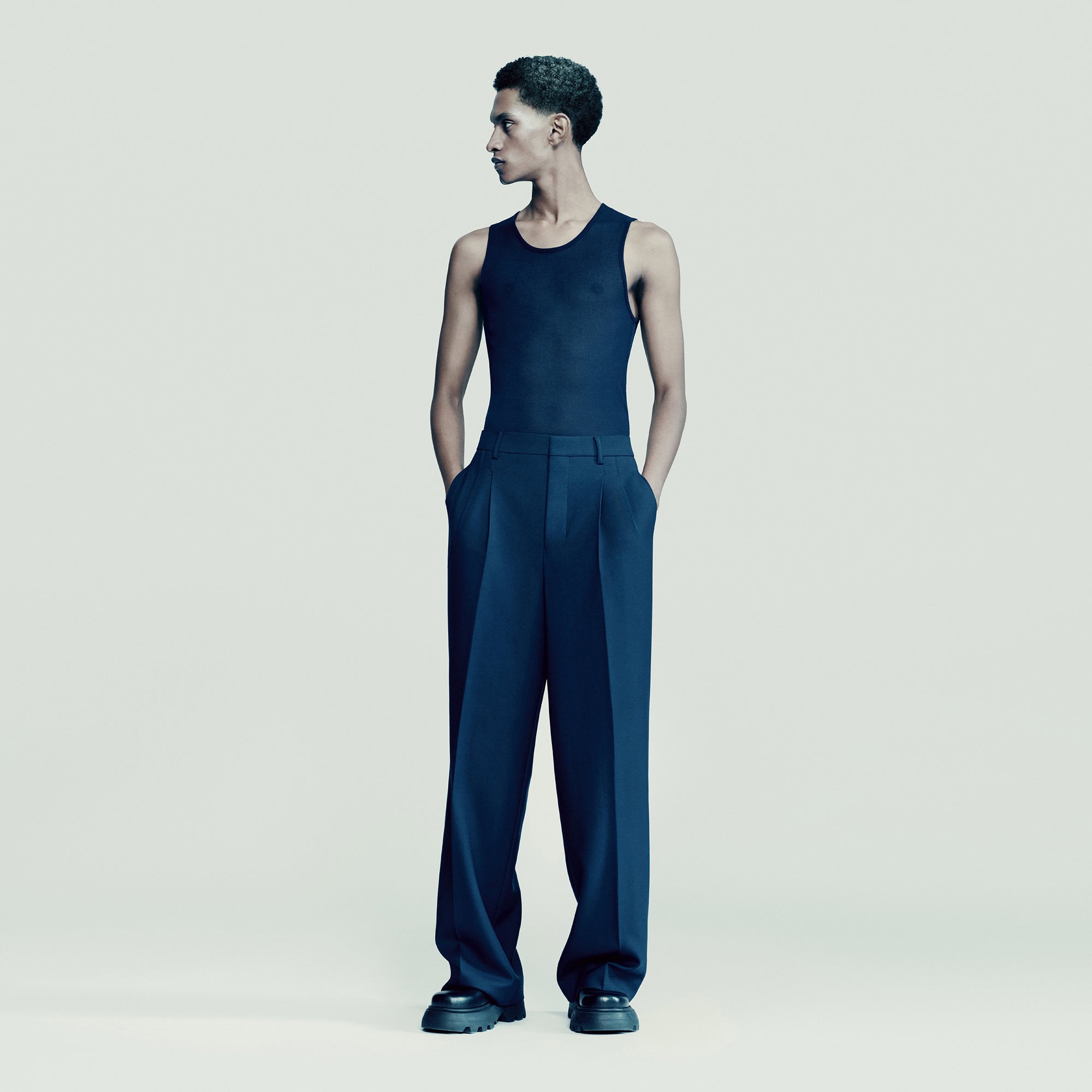 Full-length studio shot of adult male model with hands in pockets, wearing a dark navy tank top and wide-legged pants against a neutral backdrop.