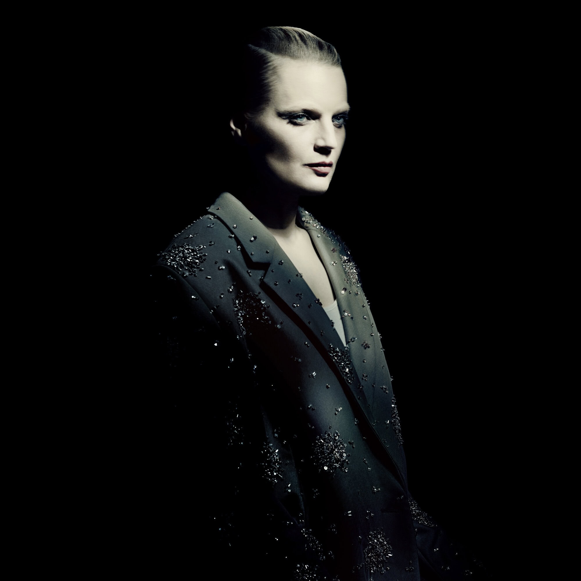 Woman poses in a dimly lit photo studio wearing a grey beaded jacket, looking right with dramatic lighting and dark background.