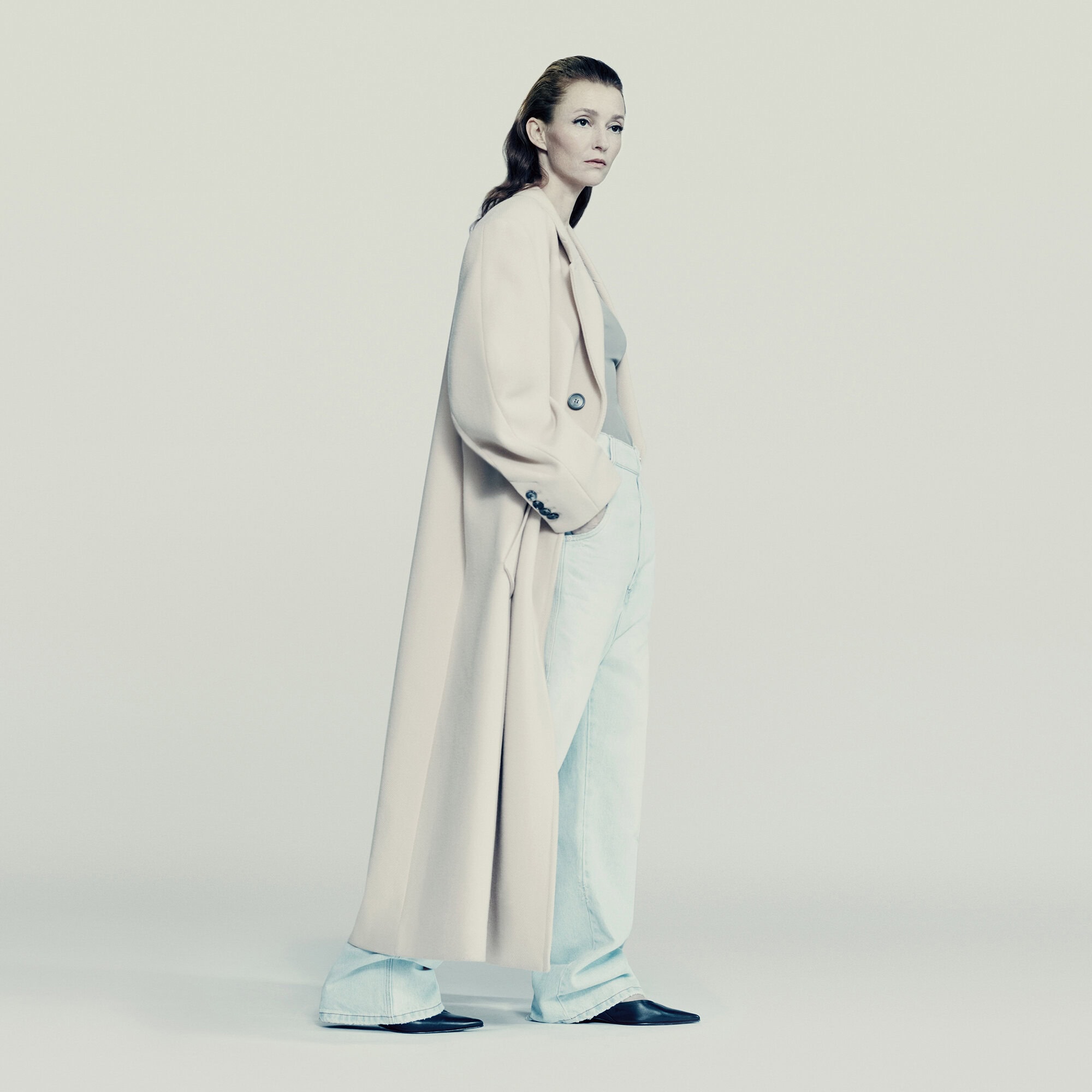 Elegant woman in a long beige coat and light blue jeans, standing indoors against a neutral backdrop in a fashion editorial shot.