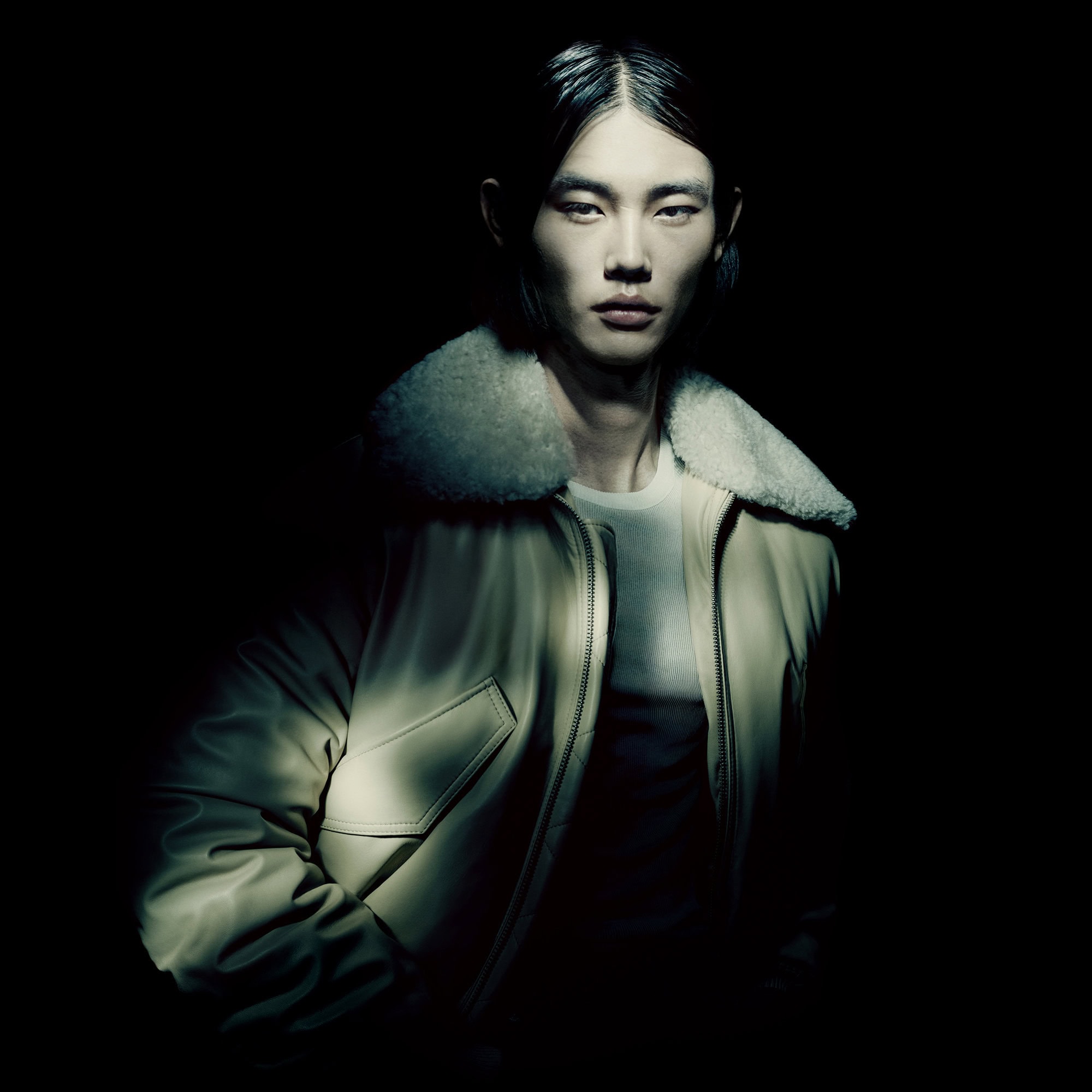 Portrait of a slender adult male with long hair posing in a creamy bomber jacket with a shearling collar against a dark backdrop.