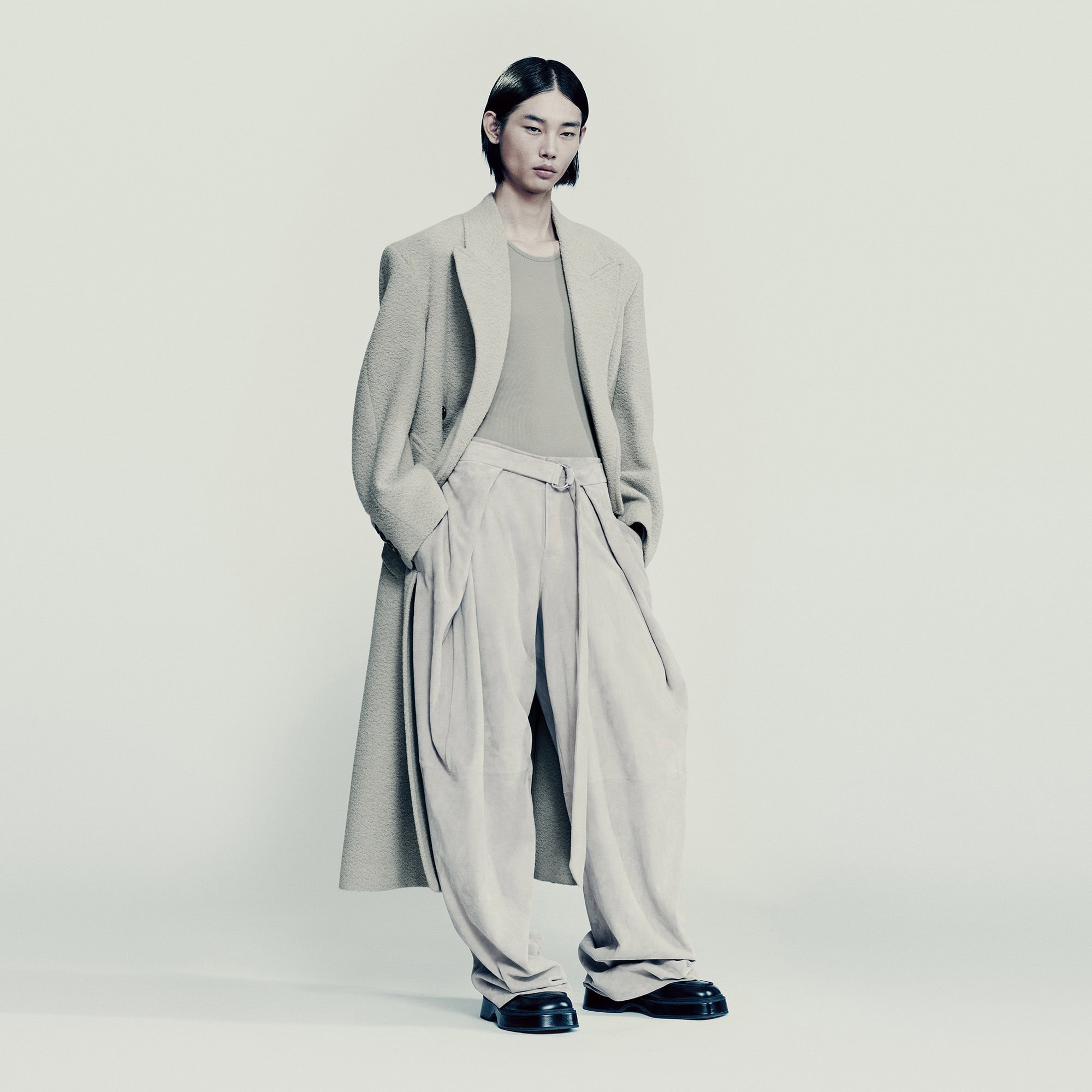 An Asian male model stands in a photo studio wearing a beige overcoat and light-beige, wide-legged pants and black leather shoes.