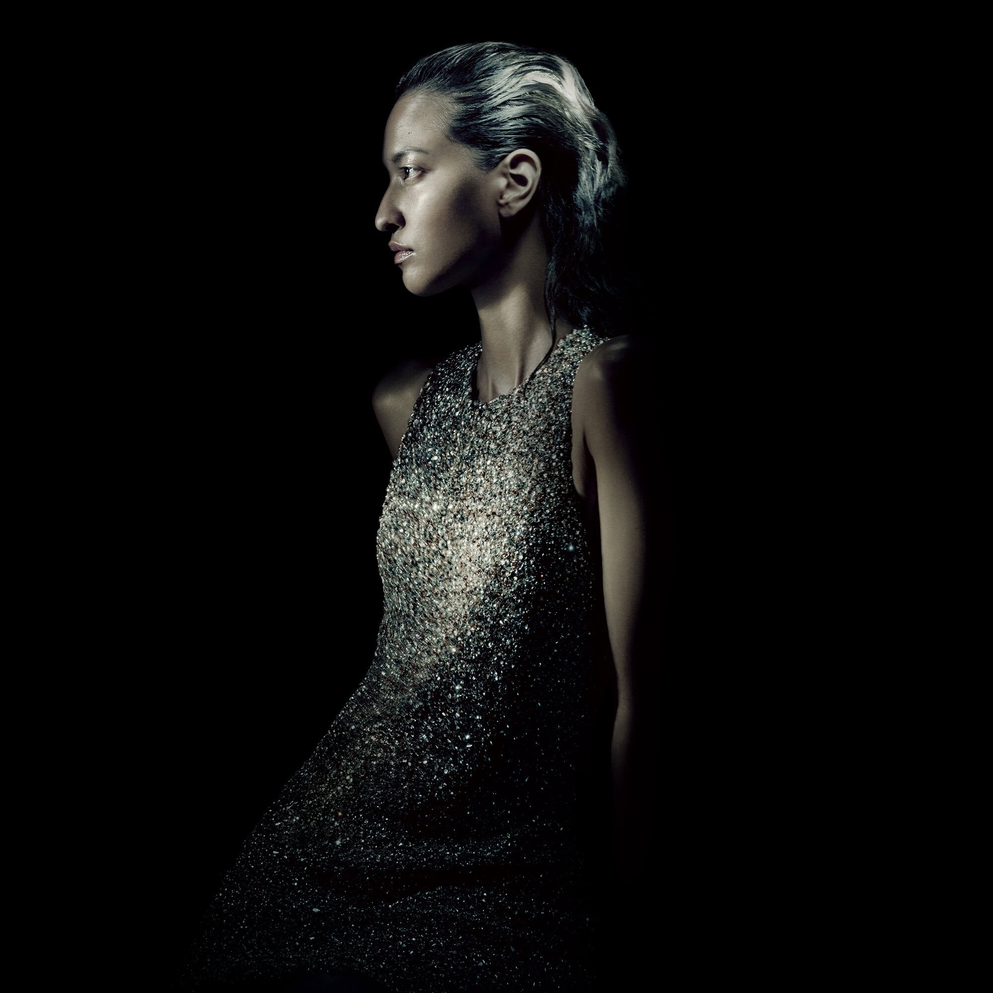 A profile view of a woman with wet-look hair wearing a silver sequin dress against a pitch-black studio background with dramatic lighting.