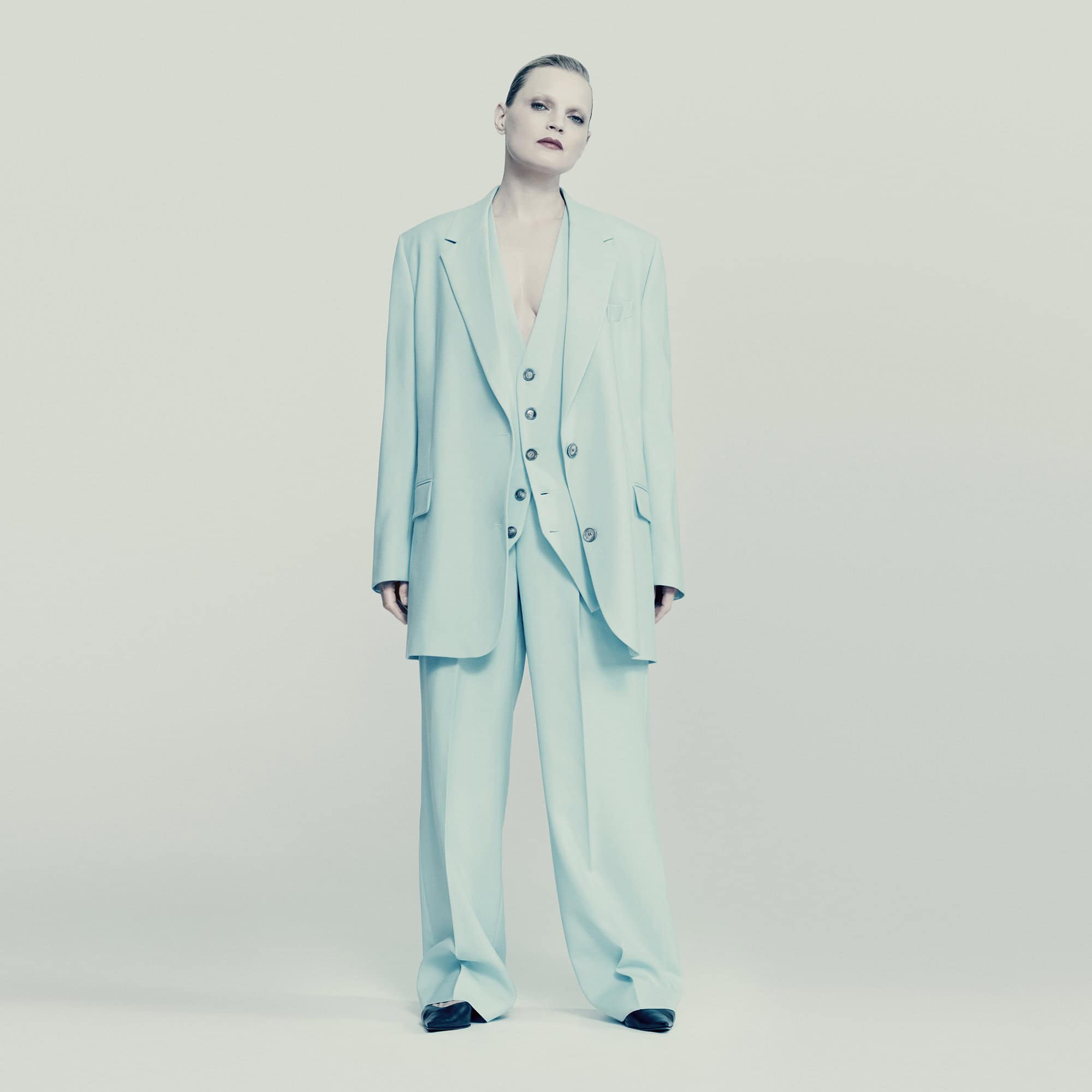 A full-length portrait of a single adult female in a light-blue pantsuit with a matching vest, standing in a light-grey studio setting.