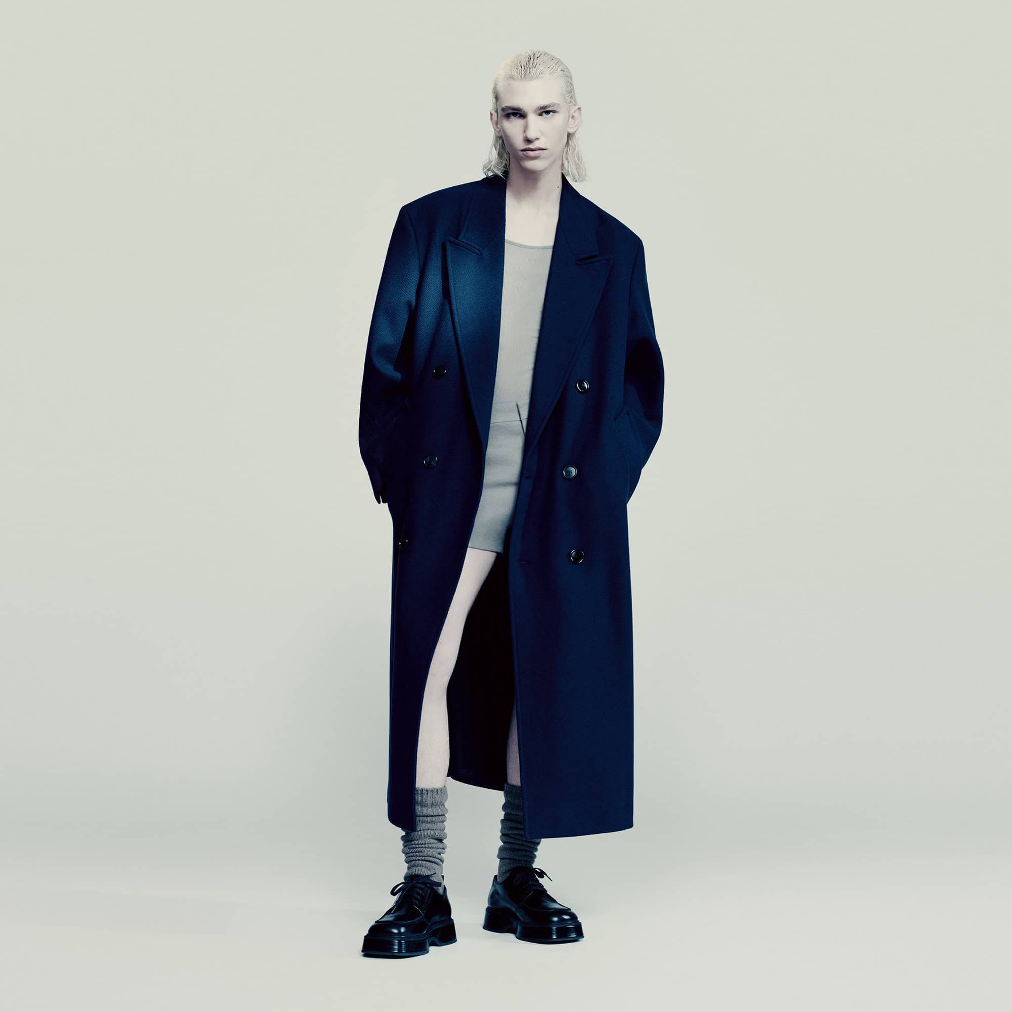 A stylish fashion shot featuring a slender person with blonde hair wearing an oversized navy coat, grey tank dress, grey socks, and black shoes.