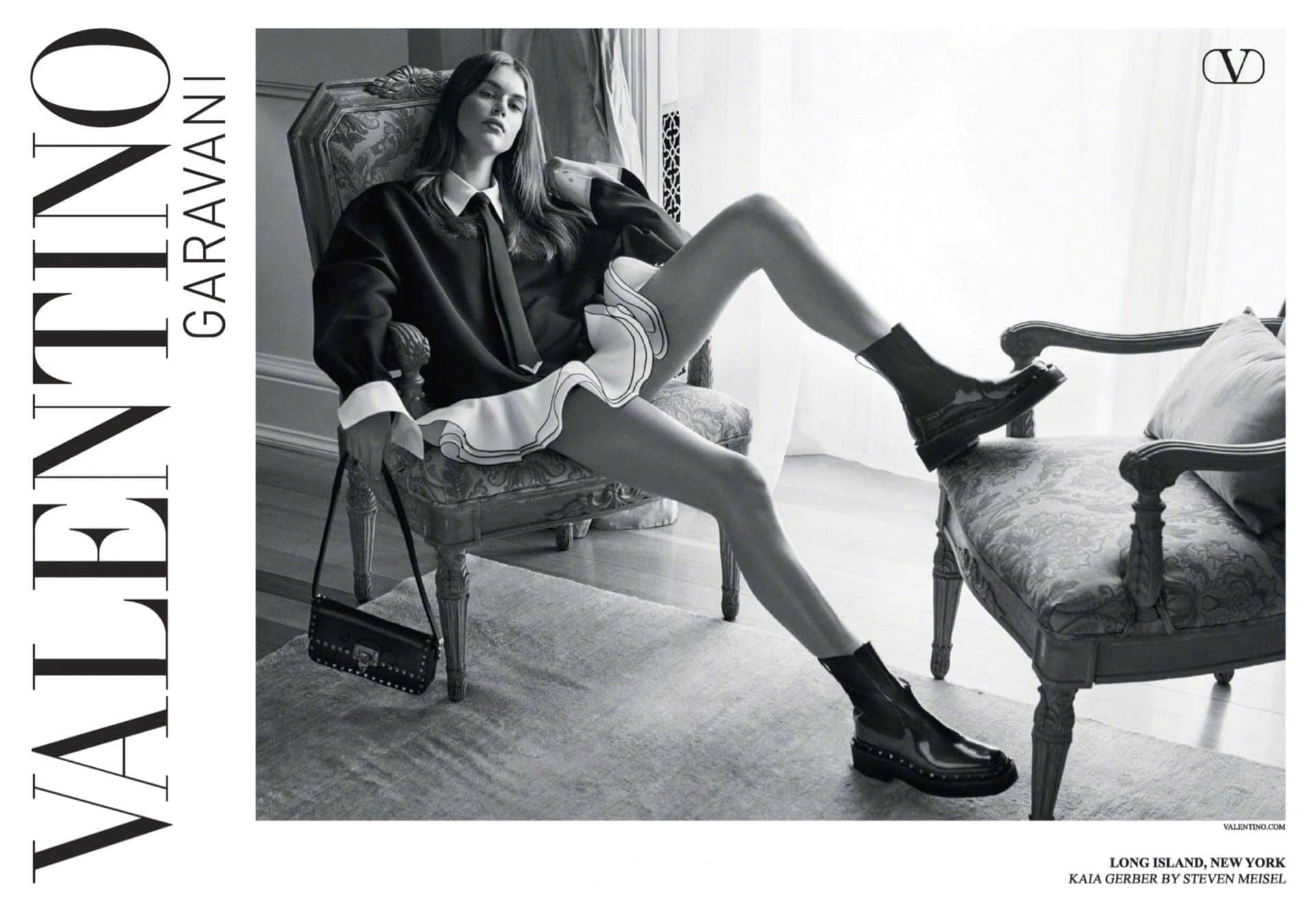 Fashion shot in black and white: A woman in a dark top and light skirt sits in an ornate chair with boots and a purse.