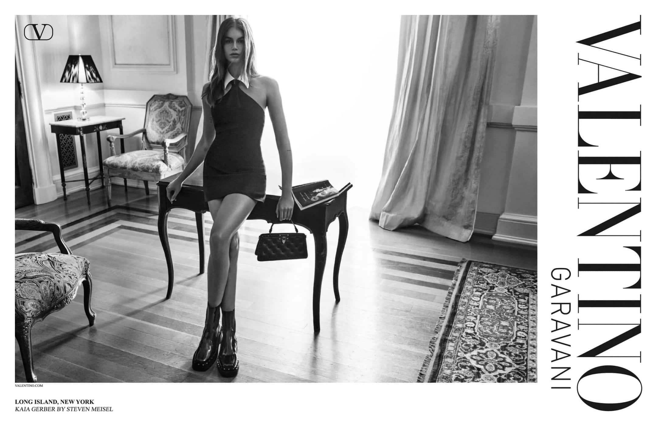 Elegant black-and-white fashion portrait of a young woman in a minidress and boots, holding a purse in an ornate interior.