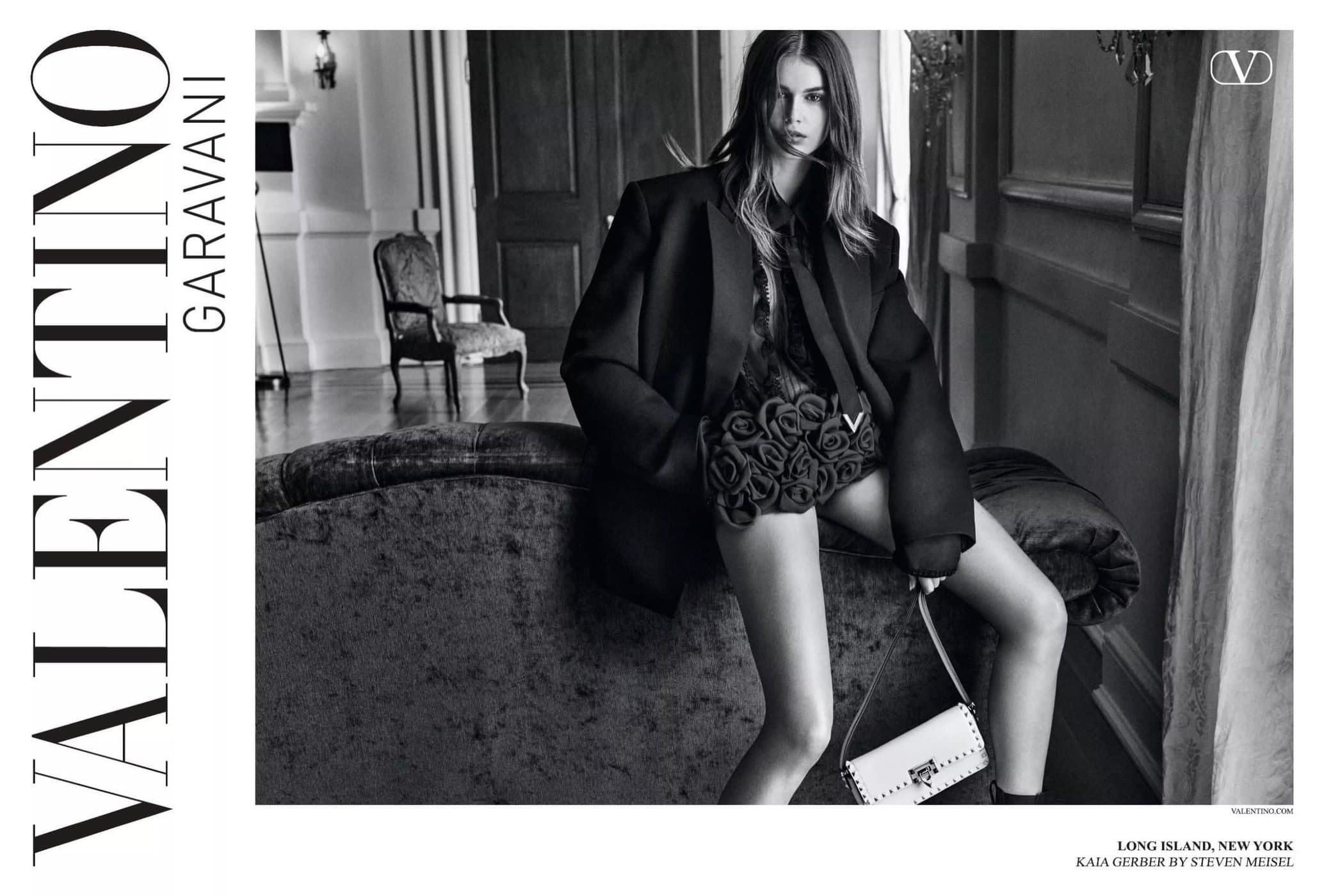Fashion shot featuring model Kaia Gerber wearing an oversized black blazer and holding a white purse, seated on a dark couch indoors.