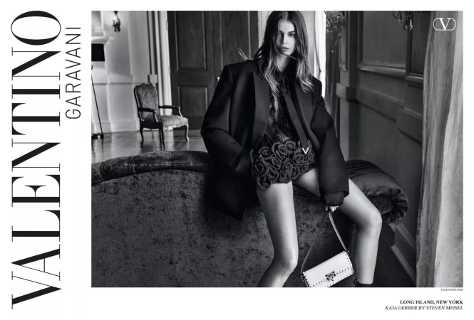 Fashion shot featuring model Kaia Gerber wearing an oversized black blazer and holding a white purse, seated on a dark couch indoors.