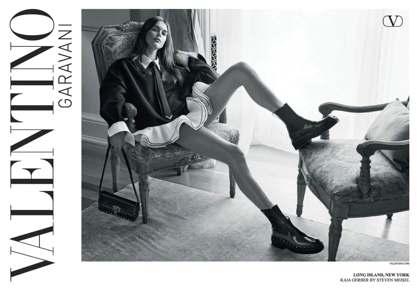 Model Kaia Gerber seated in an armchair wearing a black-and-white outfit for Valentino Black Tie Fall 2023 campaign.