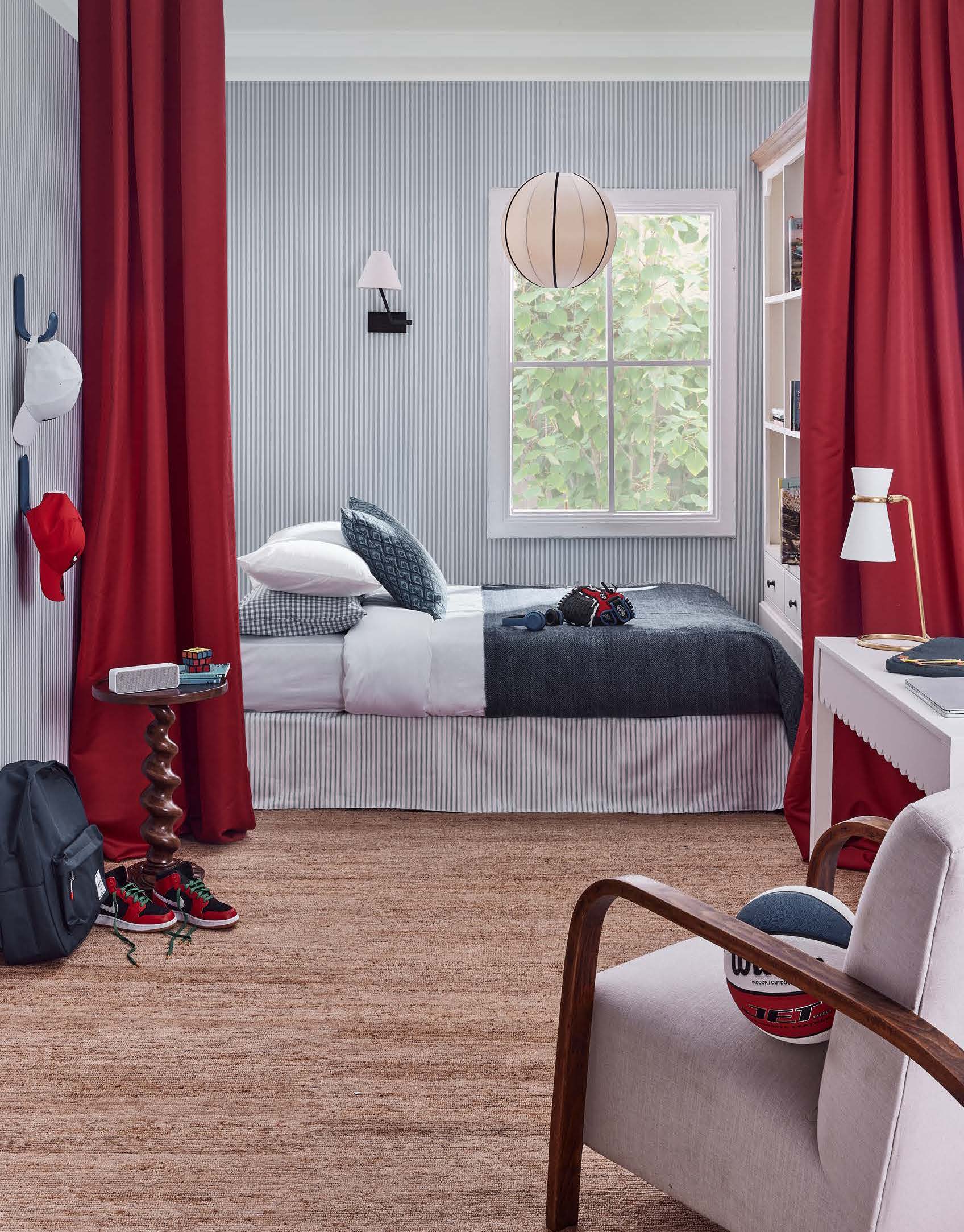 Interior shot of a child's bedroom featuring a bed, side table, chair, basketball, and red curtains against striped wallpaper.