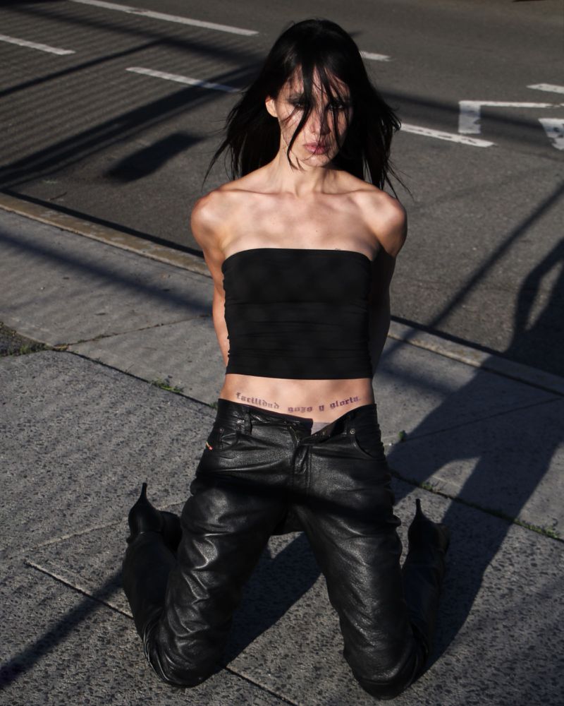 Young woman kneels on asphalt in a black strapless top and leather pants, looking at the camera with harsh sunlight casting shadows.