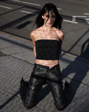 Young woman kneels on asphalt in a black strapless top and leather pants, looking at the camera with harsh sunlight casting shadows.