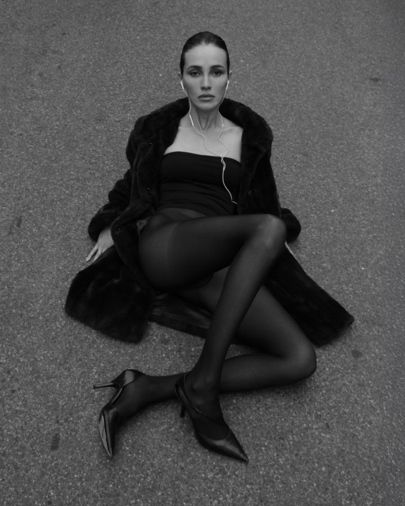 Black and white portrait of a woman sitting in a coat and heels, wearing headphones, looking directly at the camera with a neutral expression.