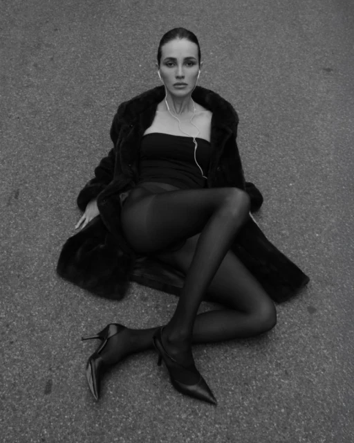 Black and white portrait of a woman sitting in a coat and heels, wearing headphones, looking directly at the camera with a neutral expression.