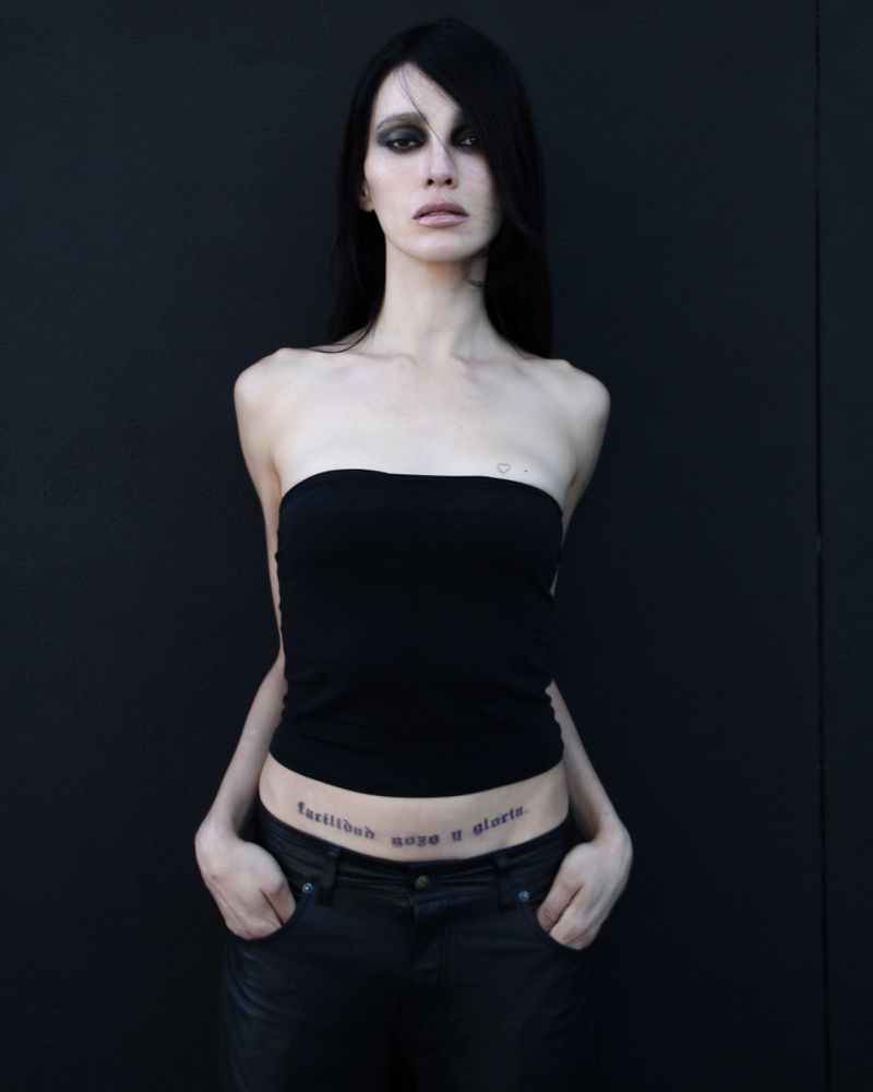 Close-up studio shot of an adult woman with dark hair wearing a black tube top and jeans, with tattoos on her abdomen, against a black backdrop.