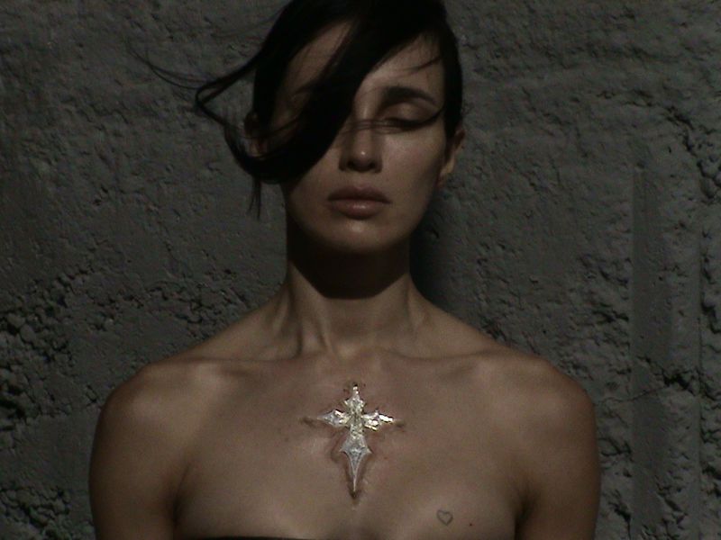 Close-up of a fair-skinned adult female with dark hair, bare shoulders, and a metallic cross pendant in front of a textured wall.