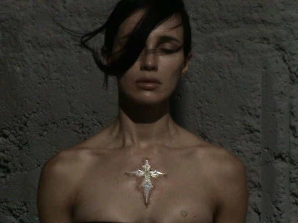 Close-up of a fair-skinned adult female with dark hair, bare shoulders, and a metallic cross pendant in front of a textured wall.