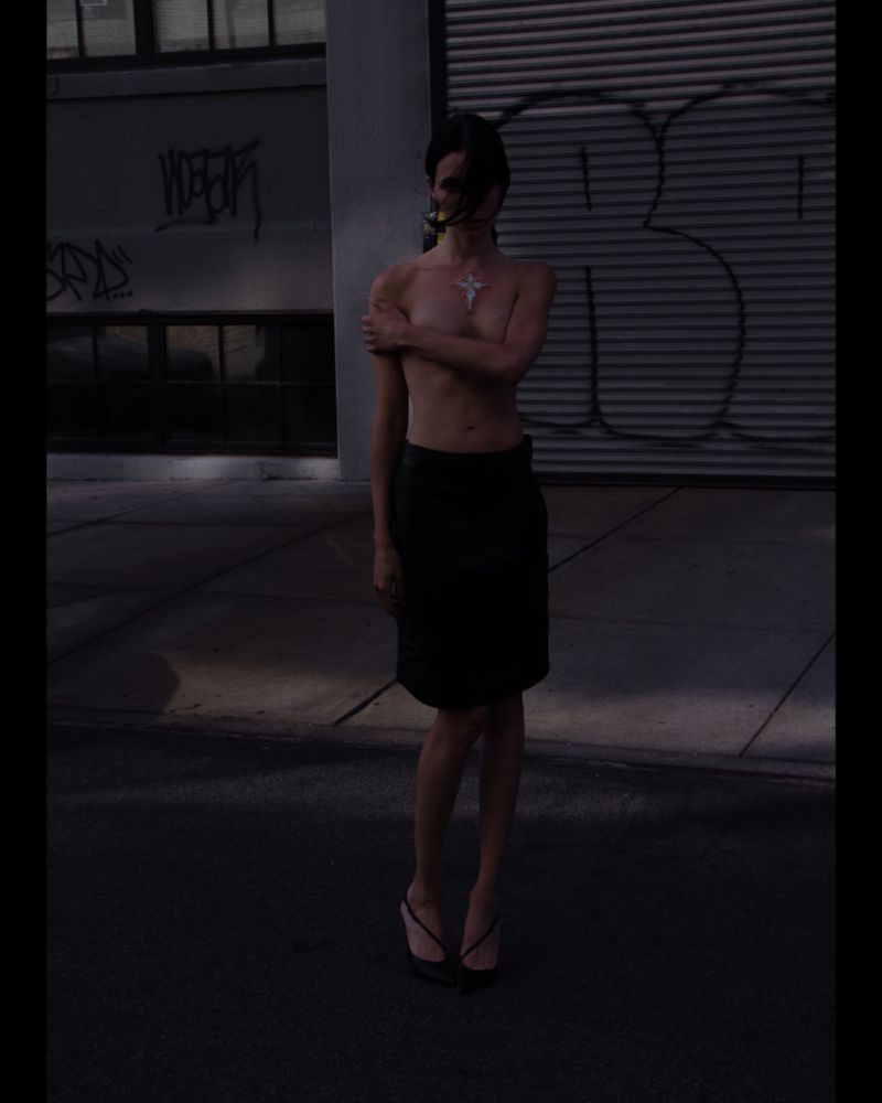 Adult female stands in street with graffiti wall in background, wearing skirt and mask. Shot in chiaroscuro style for dramatic effect.