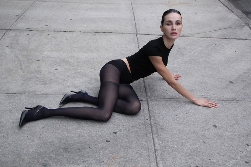 Adult female model in black top, sheer tights, and high heels lying on concrete surface in daylight. Fashion editorial style.