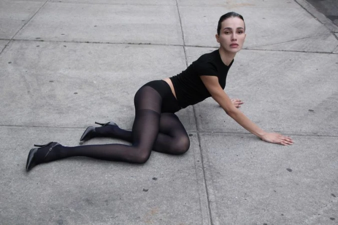 Adult female model in black top, sheer tights, and high heels lying on concrete surface in daylight. Fashion editorial style.