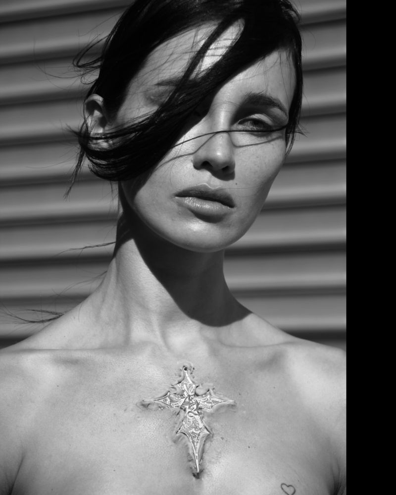Monochrome portrait of an adult woman with dark hair, fair skin, and a decorative cross pendant necklace. Outdoor setting with light shadows.